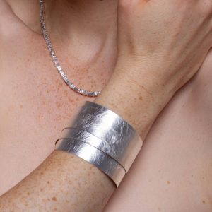 Flexibler Armreif und quadratische Halskette in Silber. Die gebürstete Oberfläche des Silberarmreifs und die quadratischen Plättchen der Silberkette spielen mit Lichtreflexen.